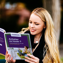 Jeune femme blonde souriante tenant un livre violet ouvert en extérieur.