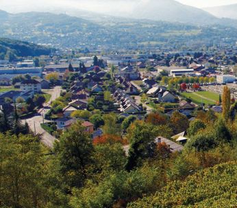 Vue panoramique d'une vallée verdoyante avec ville et montagnes enneigées en arrière-plan sous ciel bleu.