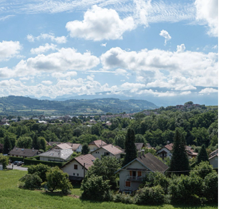 Paysage montagneux avec vallée verdoyante et maisons dispersées sous ciel nuageux.