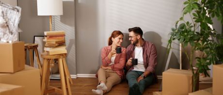 Jeune couple souriant assis sur le sol parmi des cartons de déménagement dans un appartement vide.