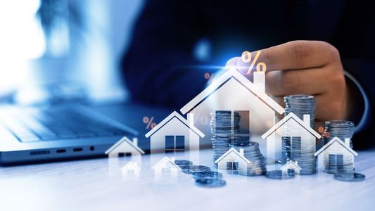 Maquette de maison blanche entourée de cubes, main la surplombant, écran numérique en arrière-plan.