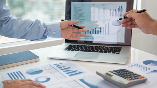 Deux personnes analysent des graphiques financiers autour d'un ordinateur portable et documents bleus sur un bureau.