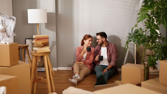Jeune couple souriant assis sur le sol parmi des cartons de déménagement dans un appartement vide.