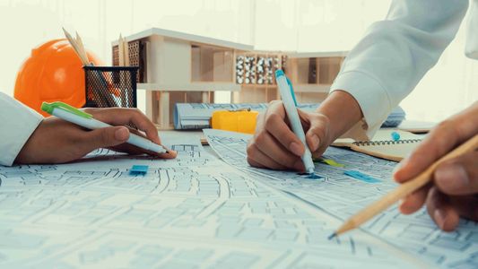 Deux architectes étudient des plans bleus sur un bureau avec casque et maquette.