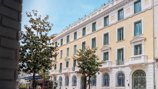 Immeuble haussmannien beige avec façade ornée et arcades au rez-de-chaussée, arbres devant.