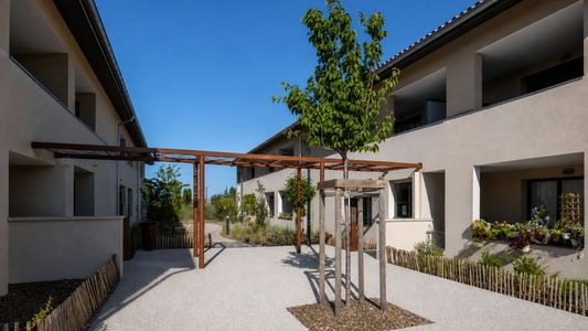 Cour intérieure avec bâtiments modernes, arbres et pavage ensoleillé sous ciel bleu.