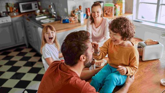 Enfants assis en cercle dans une cuisine rétro, levant les mains vers une adulte souriante.