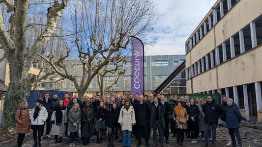 Un groupe de personnes rassemblées dans une cour urbaine ensoleillée, entourées d'arbres nus et de bâtiments.