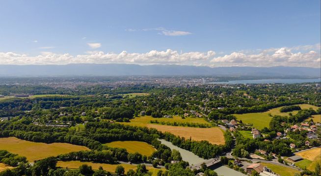 Vue aérienne d'une vallée verdoyante avec champs jaunes, rivière sinueuse et villages sous ciel bleu nuageux.