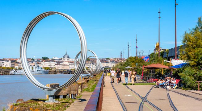 Grande roue argentée au bord de l'eau avec promenade et installations colorées sous ciel bleu dégagé.