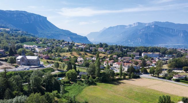 Vue aérienne d'une vallée alpine avec montagnes boisées, villages dispersés et champs verts.