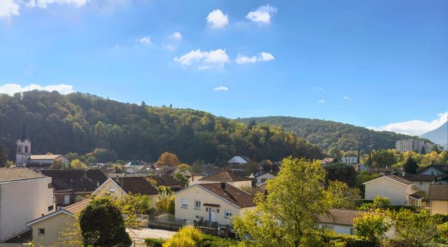 Vue aérienne d'un village vallonné avec maisons dispersées parmi la verdure sous un ciel bleu dégagé.