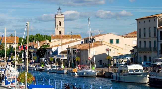 Port méditerranéen avec bateaux, église campanile et bâtiments colorés sous ciel bleu.