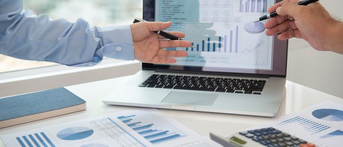 Deux personnes analysent des graphiques financiers autour d'un ordinateur portable et documents bleus sur un bureau.