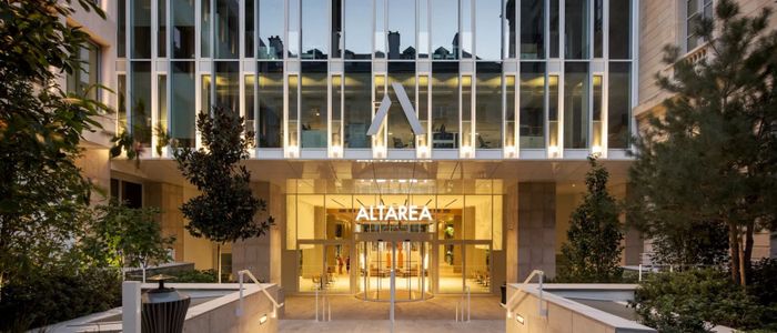 Entrée illuminée d'un bâtiment moderne avec colonnes blanches, flanquée de plantes vertes et paroi vitrée dorée.