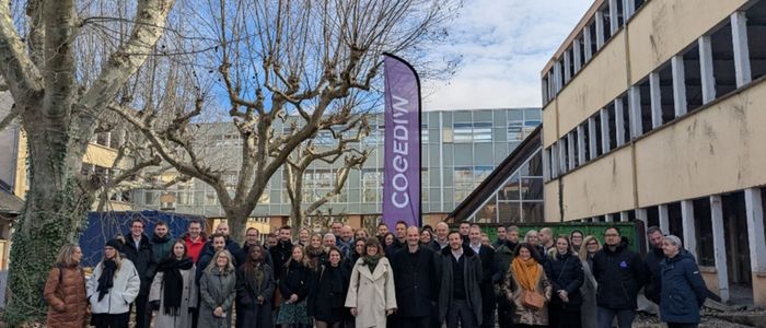 Un groupe de personnes rassemblées dans une cour urbaine ensoleillée, entourées d'arbres nus et de bâtiments.